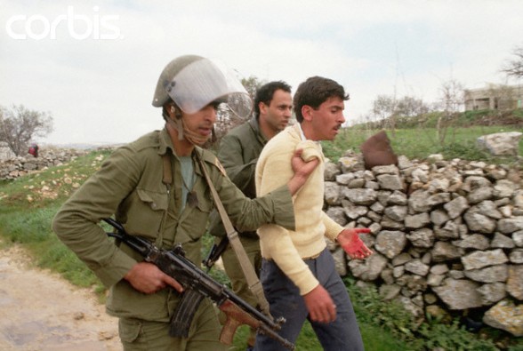 ca. March 1988, West Bank --- Two Israeli soldiers force a bleeding Palestinian man to walk with them down an unpaved road. --- Image by  Peter Turnley/CORBIS